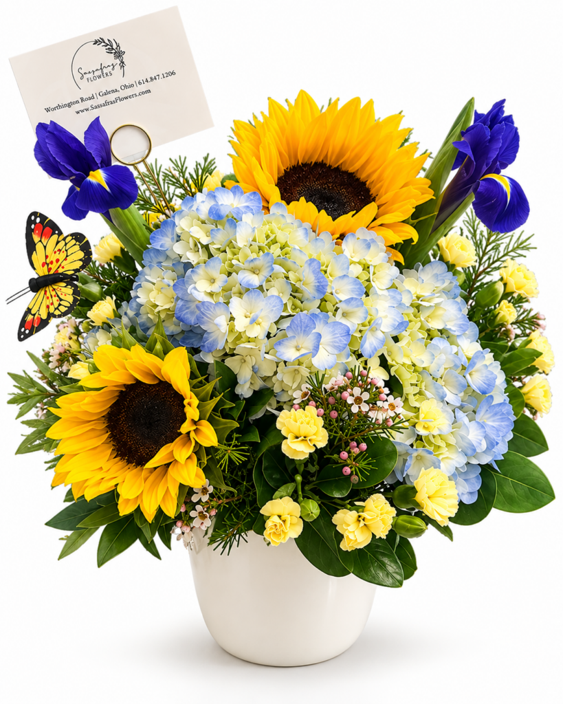 A Little Sunshine floral arrangement featuring yellow sunflowers, blue hydrangeas, purple irises, and seasonal blooms with a butterfly accent in a white ceramic vase by Sassafraz Flowers, Galena, Ohio.