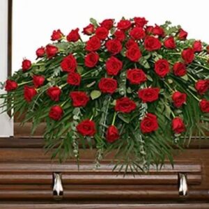 True Love's Tribute red rose casket spray featuring abundant red roses and cascading greenery displayed atop a wooden casket by Sassafraz Flowers, Galena, Ohio.