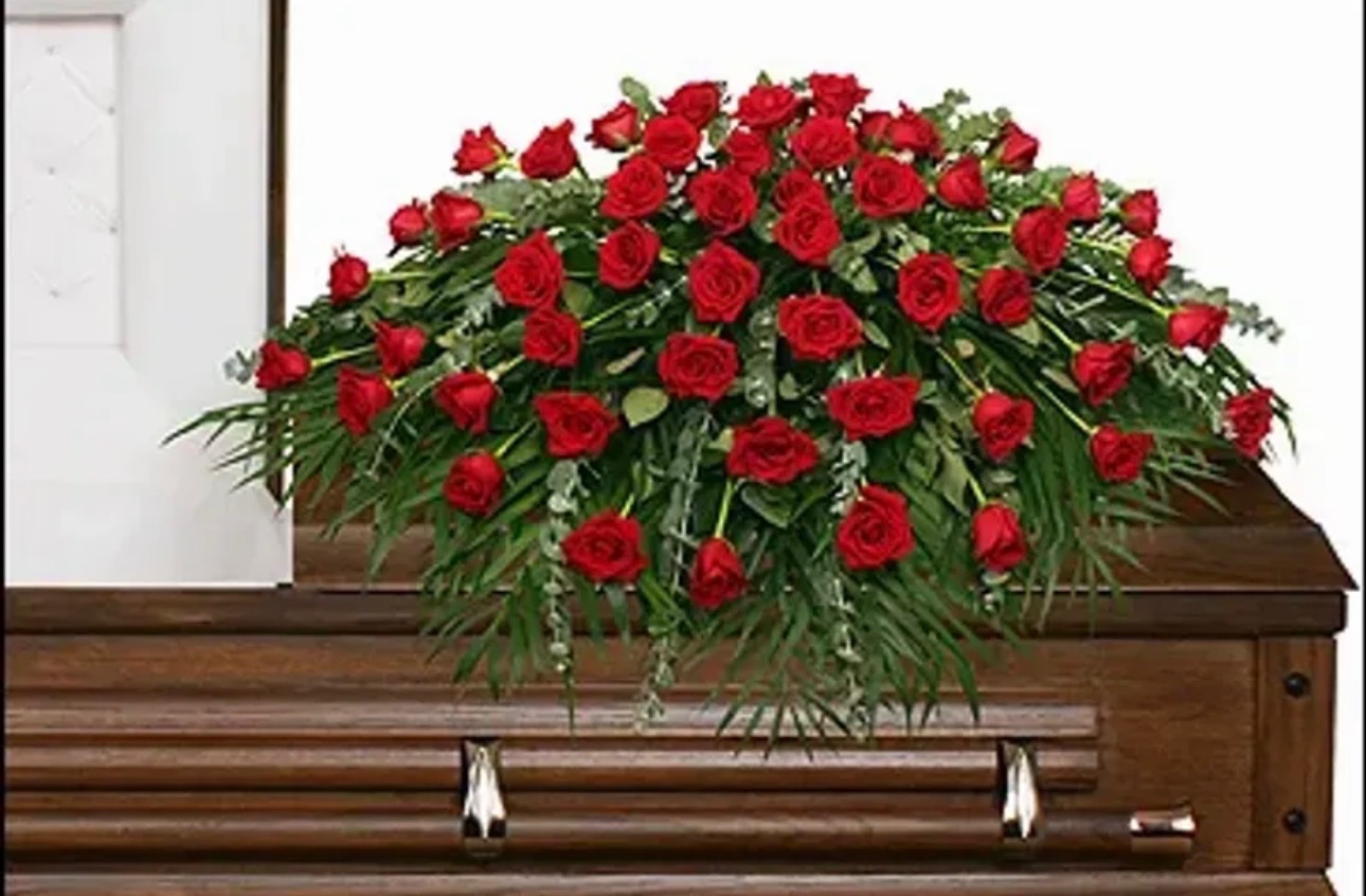 True Love's Tribute red rose casket spray featuring abundant red roses and cascading greenery displayed atop a wooden casket by Sassafraz Flowers, Galena, Ohio.