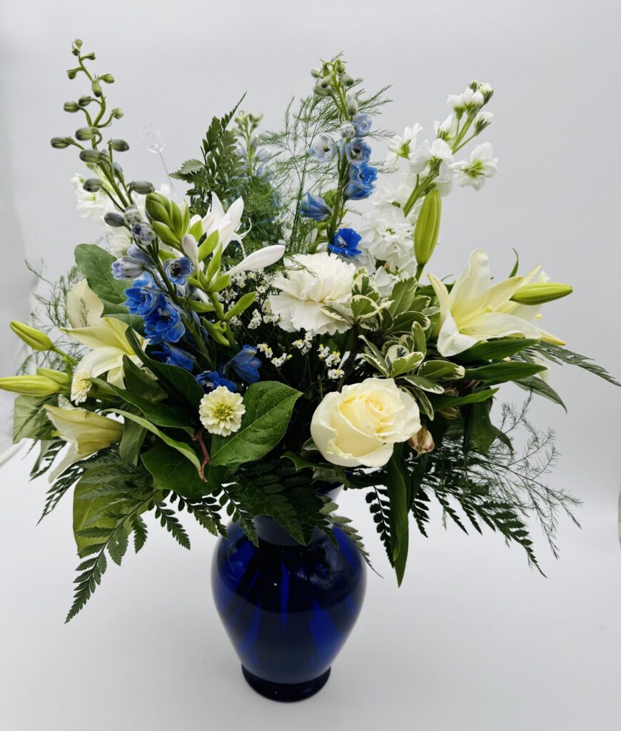 Clear Reflection of the Sky floral arrangement featuring white lilies, ivory roses, blue delphinium, and lush greenery in a cobalt blue glass vase by Sassafraz Flowers, Galena, Ohio.