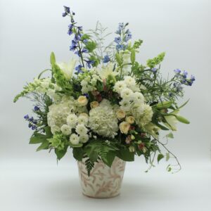Inspired by the quiet beauty of nature's renewal, First Day of Spring is a graceful celebration of fresh beginnings, delicate color, and effortless elegance. Abundant white hydrangeas create a soft, cloudlike foundation, complemented by creamy ivory roses, elegant white lilies, and delicate blue delphinium. Layers of seasonal blossoms, airy botanical textures, and naturally flowing greenery introduce movement and dimension, creating a composition that feels beautifully organic and thoughtfully composed. Presented in a decorative ceramic vessel with subtle botanical detailing, First Day of Spring balances classic floral beauty with a fresh, garden-inspired sensibility. Its generous proportions and graceful silhouette make it a distinctive statement for a home, office, or special gathering. Whether celebrating a birthday, welcoming a new beginning, expressing appreciation, or offering comfort and encouragement, First Day of Spring brings a sense of optimism, tranquility, and natural beauty to life's meaningful moments. Each arrangement is individually handcrafted with care. Seasonal flowers, greenery, and vessels may vary while preserving the overall color palette, character, and quality of the design.
