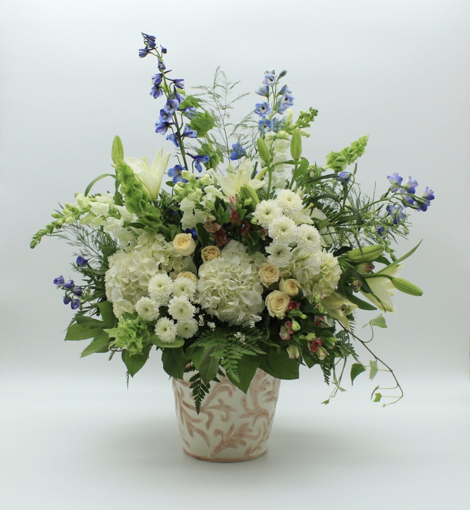 Inspired by the quiet beauty of nature's renewal, First Day of Spring is a graceful celebration of fresh beginnings, delicate color, and effortless elegance. Abundant white hydrangeas create a soft, cloudlike foundation, complemented by creamy ivory roses, elegant white lilies, and delicate blue delphinium. Layers of seasonal blossoms, airy botanical textures, and naturally flowing greenery introduce movement and dimension, creating a composition that feels beautifully organic and thoughtfully composed. Presented in a decorative ceramic vessel with subtle botanical detailing, First Day of Spring balances classic floral beauty with a fresh, garden-inspired sensibility. Its generous proportions and graceful silhouette make it a distinctive statement for a home, office, or special gathering. Whether celebrating a birthday, welcoming a new beginning, expressing appreciation, or offering comfort and encouragement, First Day of Spring brings a sense of optimism, tranquility, and natural beauty to life's meaningful moments. Each arrangement is individually handcrafted with care. Seasonal flowers, greenery, and vessels may vary while preserving the overall color palette, character, and quality of the design.