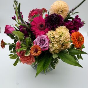 Garden Party floral arrangement featuring pink gerbera daisies, lavender roses, peach dahlias, orange zinnias, hydrangeas, and lush greenery in a glass vase by Sassafraz Flowers, Galena, Ohio.