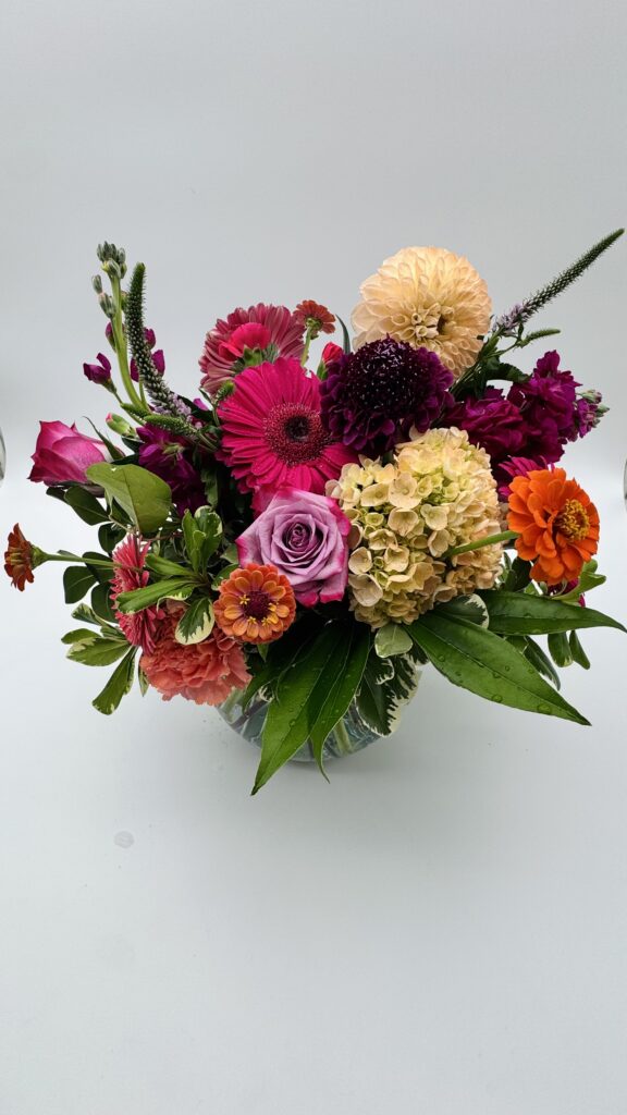 Garden Party floral arrangement featuring pink gerbera daisies, lavender roses, peach dahlias, orange zinnias, hydrangeas, and lush greenery in a glass vase by Sassafraz Flowers, Galena, Ohio.