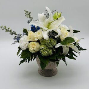 Pure Grace floral arrangement featuring white lilies, ivory roses, white carnations, green hydrangeas, and delicate blue accents in a decorative ceramic vase by Sassafraz Flowers, Galena, Ohio.