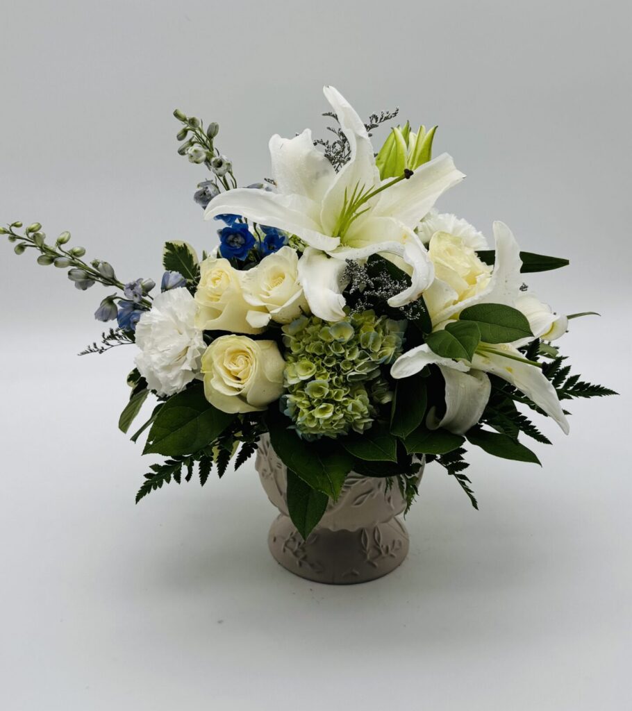 Pure Grace floral arrangement featuring white lilies, ivory roses, white carnations, green hydrangeas, and delicate blue accents in a decorative ceramic vase by Sassafraz Flowers, Galena, Ohio.