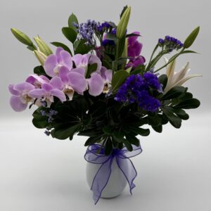Purple Vineyard floral arrangement featuring lavender orchids, purple seasonal blooms, white lily buds, and lush greenery in a white ceramic vase with a purple ribbon by Sassafraz Flowers, Galena, Ohio.