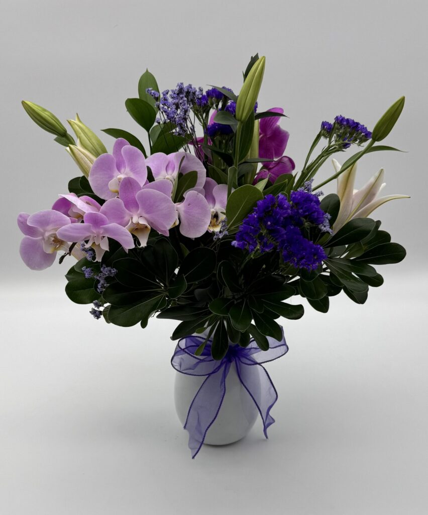 Purple Vineyard floral arrangement featuring lavender orchids, purple seasonal blooms, white lily buds, and lush greenery in a white ceramic vase with a purple ribbon by Sassafraz Flowers, Galena, Ohio.
