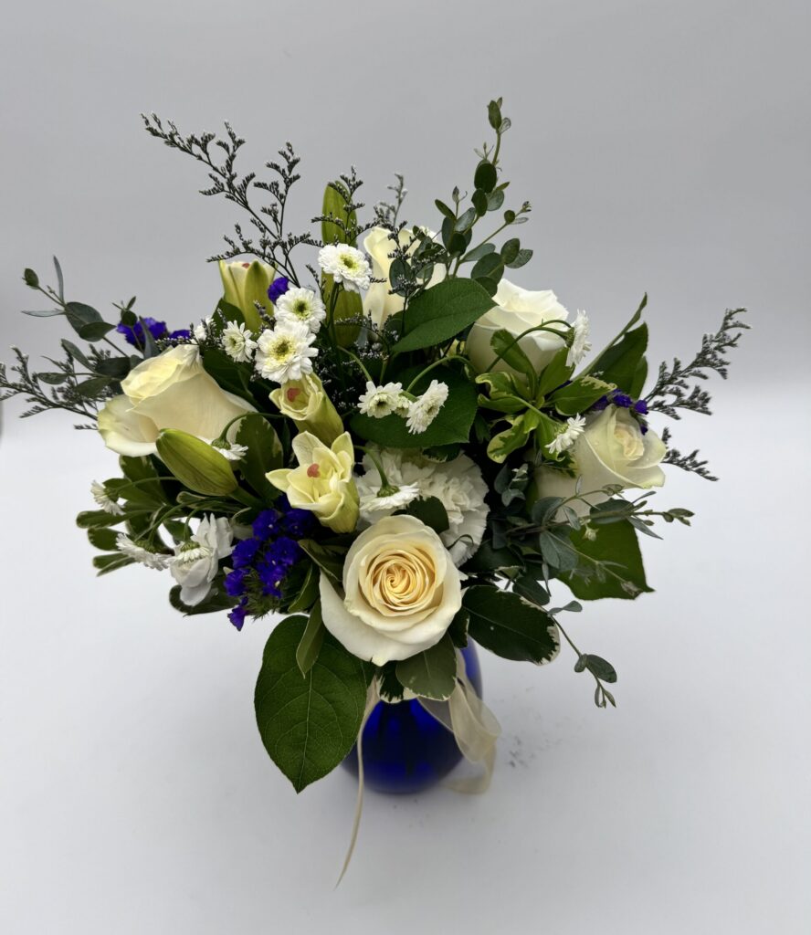 Reflections floral arrangement featuring ivory roses, white seasonal blooms, deep blue accents, and greenery in a cobalt blue vase by Sassafraz Flowers, Galena, Ohio.