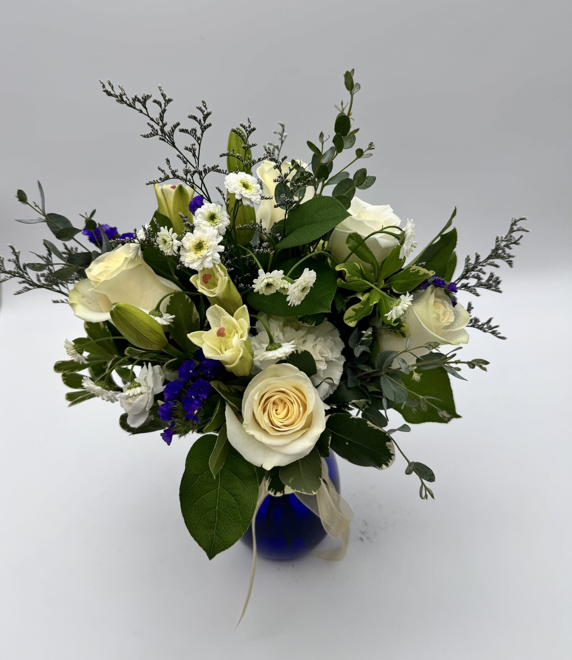 Reflections floral arrangement featuring ivory roses, white seasonal blooms, deep blue accents, and greenery in a cobalt blue vase by Sassafraz Flowers, Galena, Ohio.